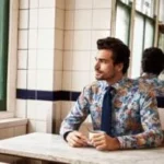 Man in a floral-patterned blazer and tie sitting at a diner counter with a coffee cup, looking out the window.