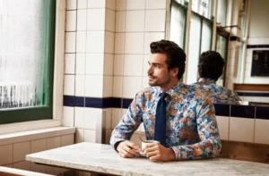 Man in a floral-patterned blazer and tie sitting at a diner counter with a coffee cup, looking out the window.