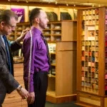 Tailor measures a customer wearing a purple dress shirt during a custom fitting in a menswear shop.