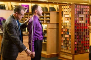 Tailor measures a customer wearing a purple dress shirt during a custom fitting in a menswear shop.