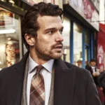Man in a tailored overcoat and plaid tie standing on a busy city street.