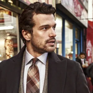 Man in a tailored overcoat and plaid tie standing on a busy city street.