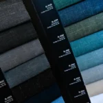A stack of dark blue, grey, and textured custom suit fabric samples labeled with design codes, displayed vertically for an upcoming tailoring event.