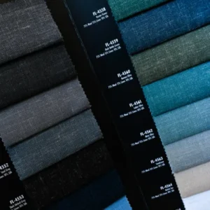 A stack of dark blue, grey, and textured custom suit fabric samples labeled with design codes, displayed vertically for an upcoming tailoring event.